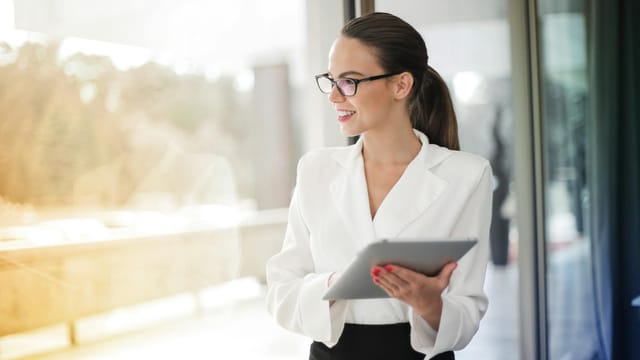 A professional woman in glasses holding a tablet in a modern office setting, exuding confidence.