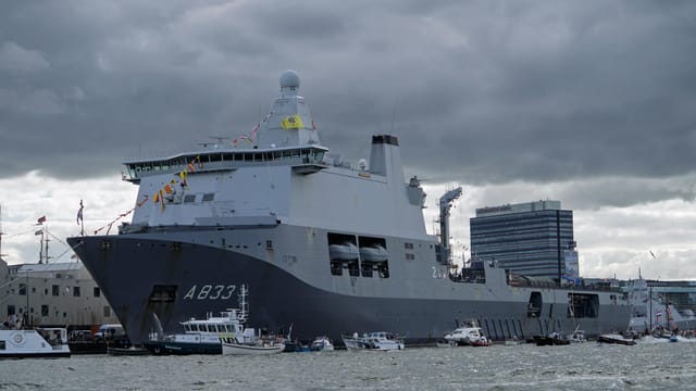 A large navy ship A833 docked in a busy harbor with smaller boats and cloudy skies.