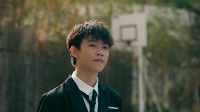A young man in formal attire smiles softly, standing in a park with a basketball hoop in the background.