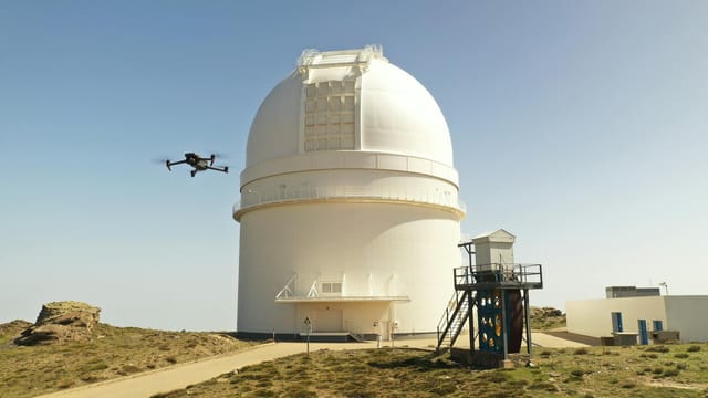A drone flies by a large observatory dome under a clear blue sky. Technology meets astronomy.