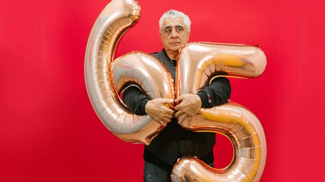 Senior man holding gold 65 number balloons against a red background.
