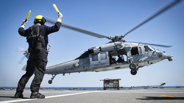 A military helicopter lands on an aircraft carrier guided by a deck crew member.