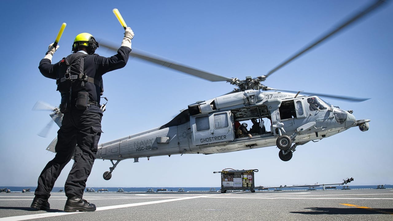 A military helicopter lands on an aircraft carrier guided by a deck crew member.