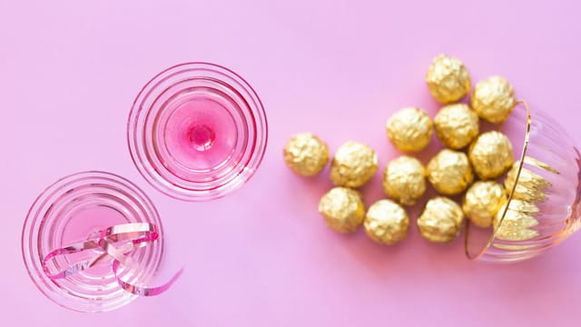 Celebrate in style with elegant champagne glasses and golden wrapped chocolates on a pink background.