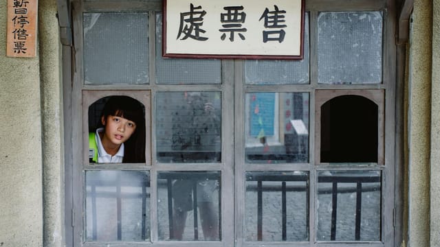 Charming ethnic teenager with focused gaze peeping out of window in building while looking at camera
