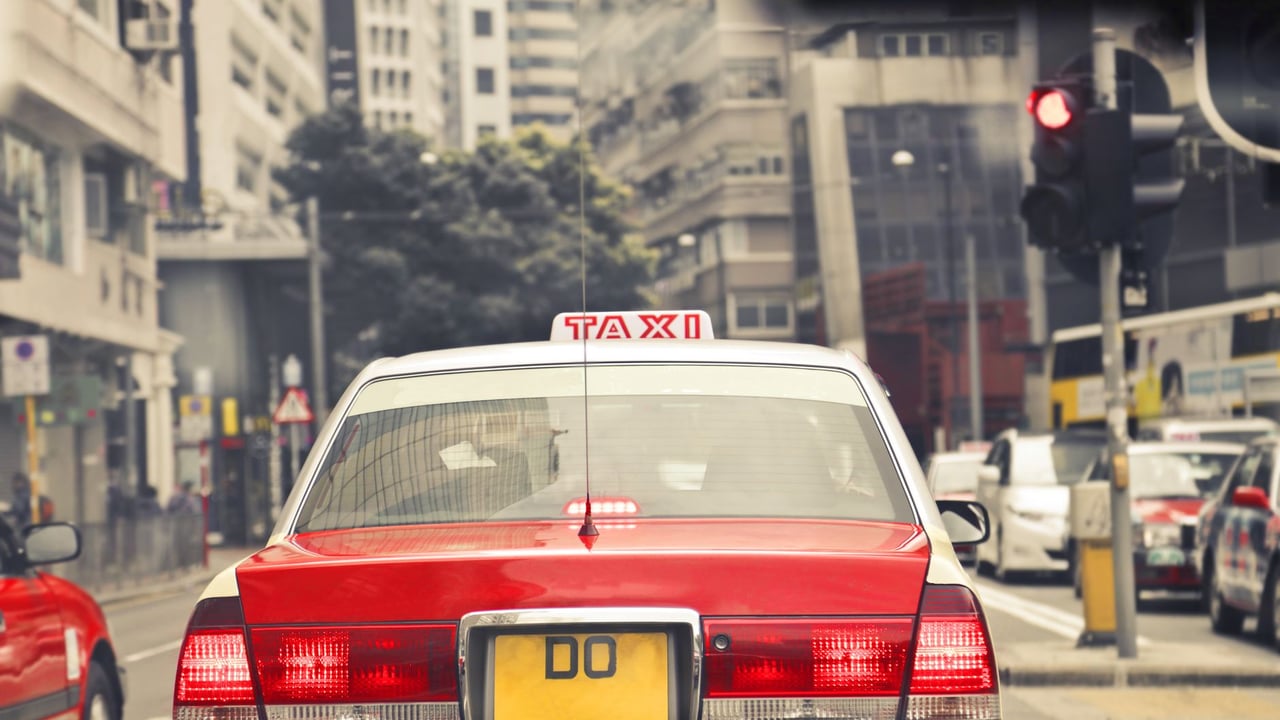Red taxi amidst city traffic on a bustling Hong Kong street, under bright daylight.