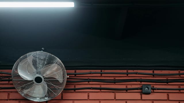 A close-up of a brick wall with a metal fan, wires, and fluorescent light. Industrial themed image.