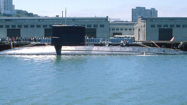 A naval submarine docked at an urban harbor with people on shore.