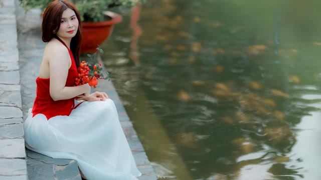 A woman in a red dress sits by a pond, holding flowers, creating a serene scene.