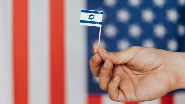 Closeup of crop unrecognizable person holding small flag of Israel before huge flag of United States of America on background