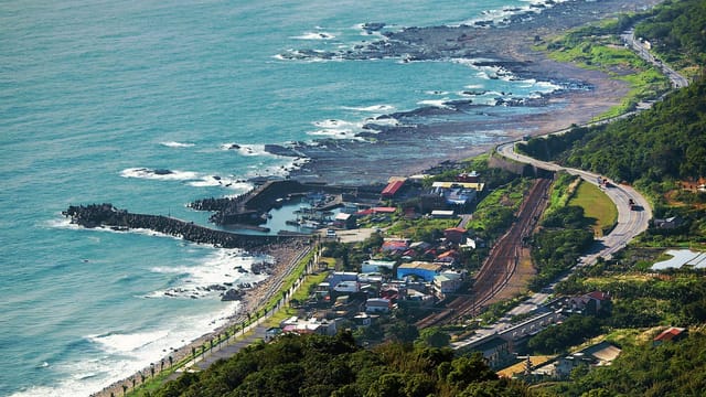 A stunning aerial shot of Taiwan's coastline, showcasing rocky shores, lush greenery, and a winding road along the ocean.