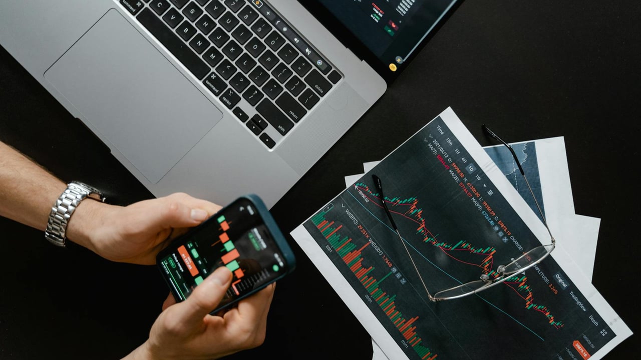 Hands using smartphone beside laptop with stock charts, showcasing digital trading.