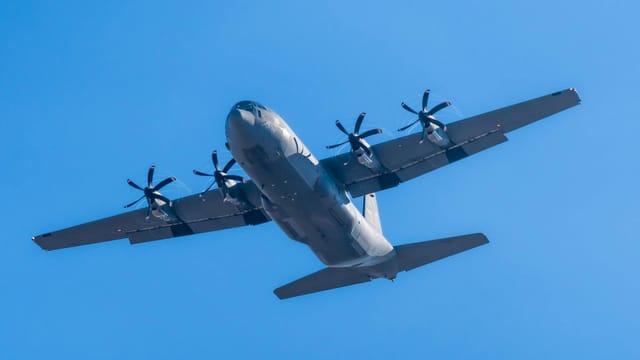 A C-130 Hercules military aircraft soaring in clear blue skies.