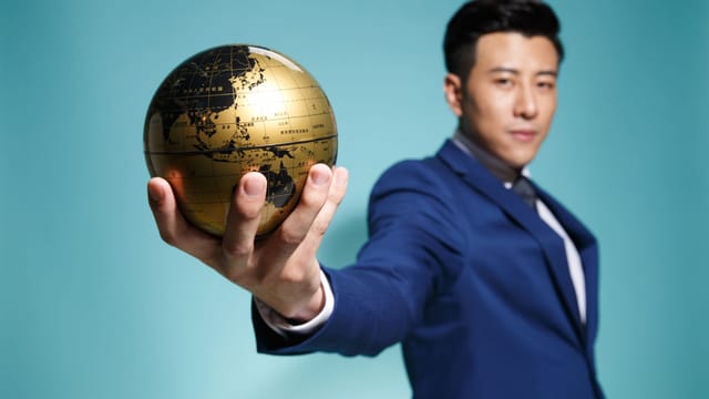 Asian businessman in blue suit confidently holding a golden globe against a blue background.