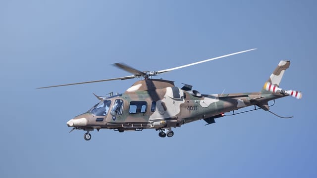 A military helicopter flies against a clear blue sky, displaying a camouflage pattern.