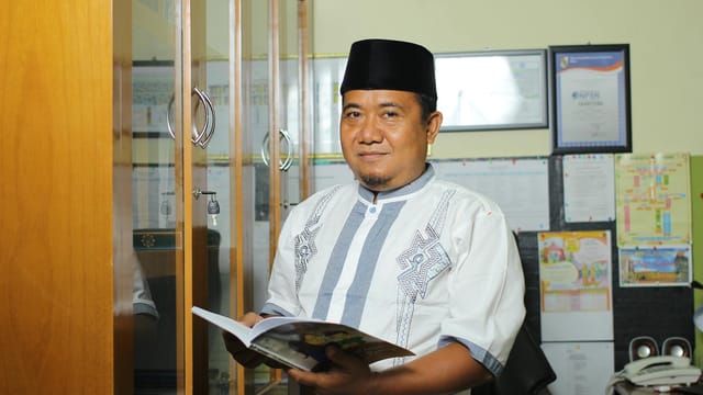 A teacher standing in an office in Indonesia, holding a book, conveying education.