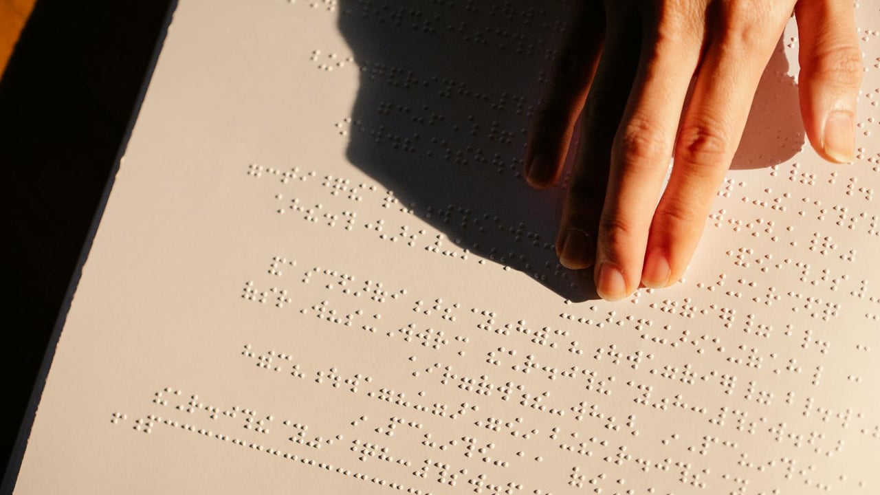 A close-up of a hand feeling and reading Braille text on paper under soft lighting.