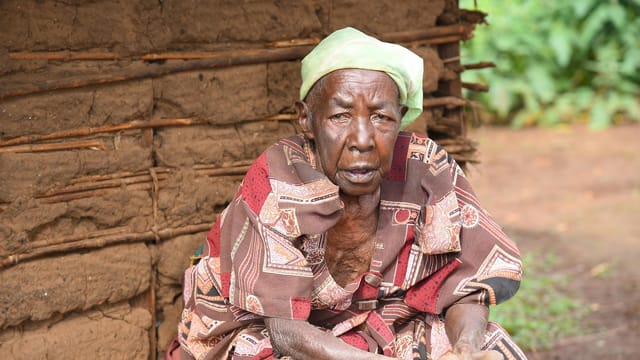 Portrait of an elderly Ugandan woman in traditional tribal attire, seated outdoors.