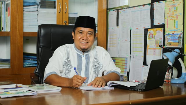 An Indonesian teacher in traditional attire, smiling at his desk with documents in an office setting.