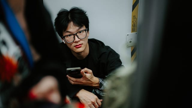 Portrait of a young man holding a smartphone indoors, looking directly at the camera.