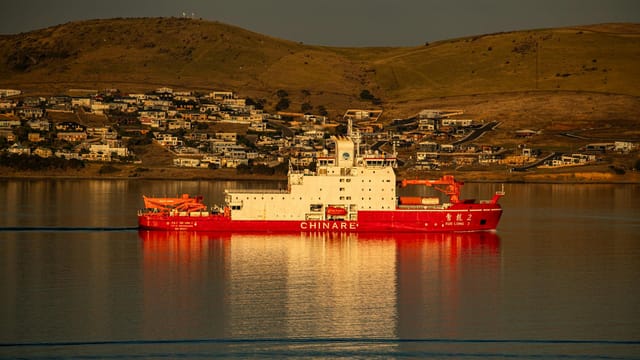 A vibrant red icebreaker ship navigating calm waters with hillside houses in the background.
