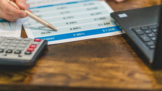 Close-up of a hand with pen analyzing financial rates on paper with a calculator and laptop nearby.
