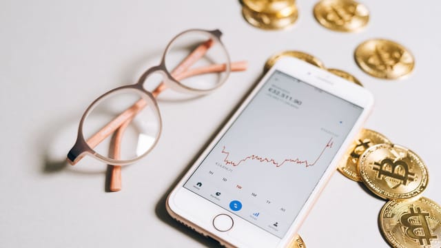 Smartphone displaying cryptocurrency graph alongside bitcoin coins and eyeglasses.