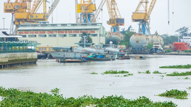 A bustling Vietnamese shipyard with cranes, military ships, and vibrant greenery.
