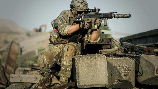 A soldier in full camouflage gear aiming a rifle while in a tactical stance outdoors.