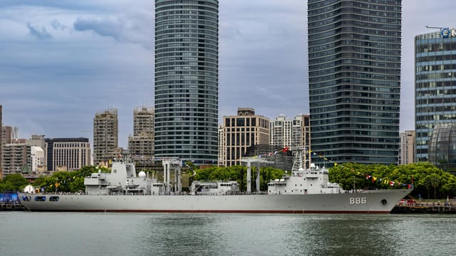 Military ship docked alongside a modern city skyline with high-rise buildings.