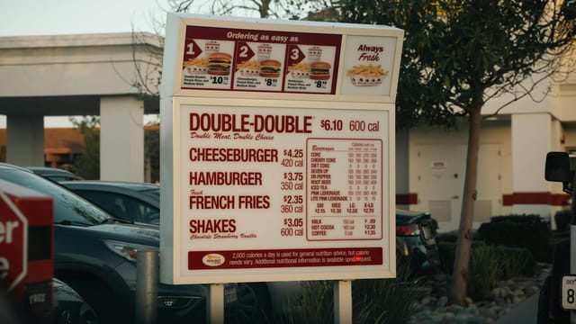 In-N-Out drive-thru menu featuring burgers, fries, and shakes under a California sky.