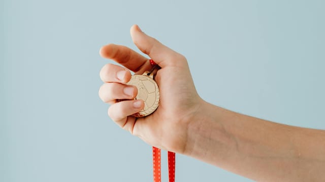 A close-up image of a hand holding a gold medal with a red ribbon against a blue background.