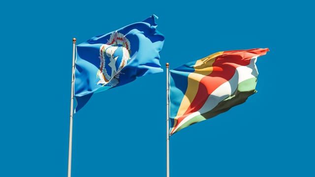 Two flags waving in the wind, representing Northern Mariana Islands and Seychelles, under a clear blue sky.