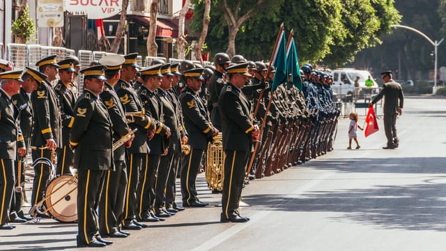 Uniformed soldiers in a ceremonial parade with Turkish flag on a city street.