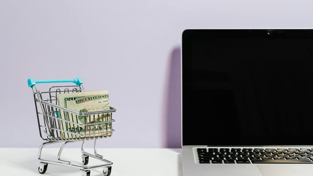Shopping cart with money next to a laptop symbolizing online shopping and e-commerce.