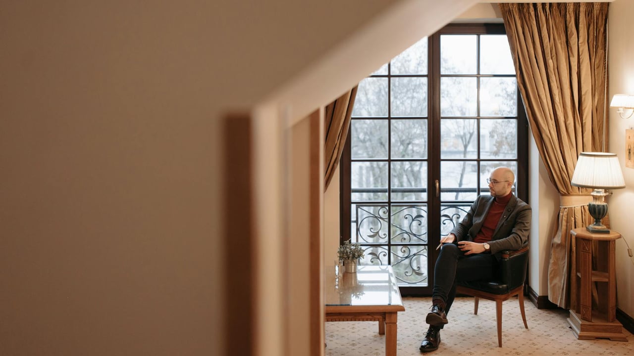 A man relaxes in a cozy living room with large windows and elegant decor.