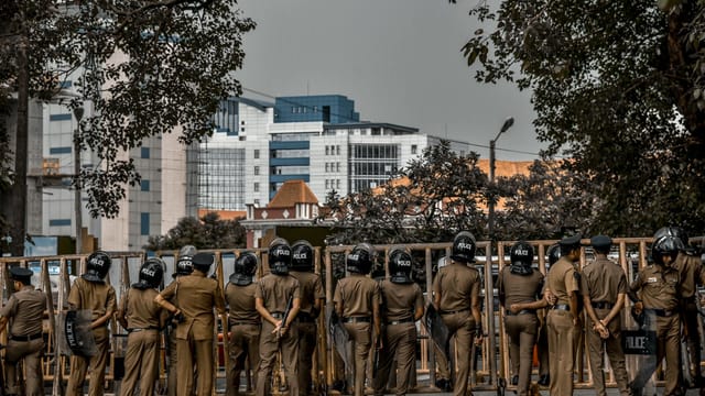 Back view of police officers in uniform standing on a city street surrounded by buildings and trees.