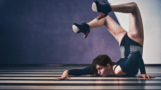 Acrobatic woman in corset and heels performing a flexible pose on a striped floor, showcasing strength and agility.