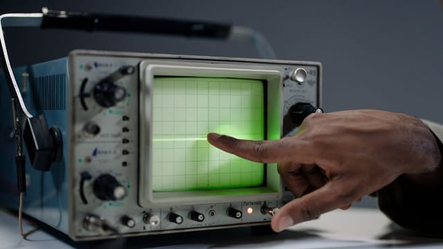 A hand pointing at the screen of a vintage oscilloscope indoors.
