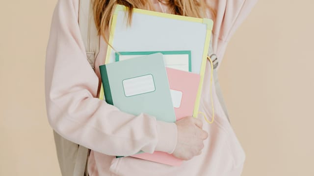 Young student in pink hoodie holding colorful notebooks, ready for school.