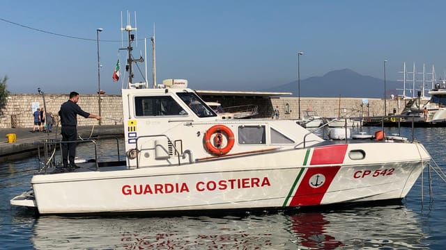 Italian Coast Guard boat docked with Mount Vesuvius in the background during a sunny day.