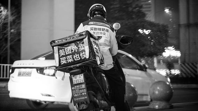 Black and white photo of a delivery driver on a scooter in urban city streets at night.