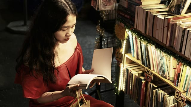 Woman in red dress reading a book at a dimly lit night market bookstall, surrounded by books.