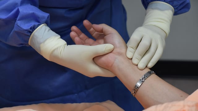 A medical professional wearing PPE checks a patient's pulse indoors.