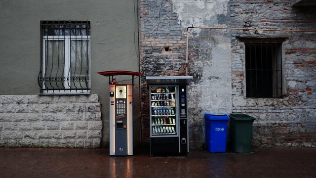 Vending machines and trash bins against an urban stone wall backdrop.