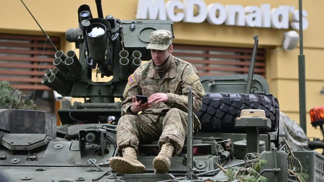 A soldier in camouflage resting on an armored vehicle, using a smartphone outside a McDonald's.