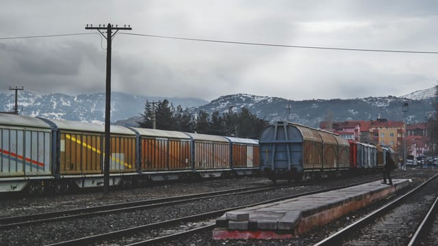 A freight train with colorful cargo wagons on a mountain railway track under a gray sky.