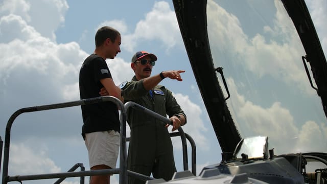 Military personnel and civilian discuss aircraft features at an airshow in Charleston, SC under clear skies.