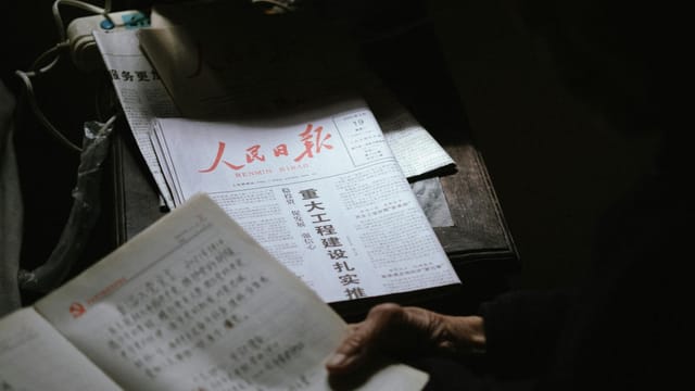 A person reading a Chinese newspaper in a dimly lit setting, creating a contemplative mood.