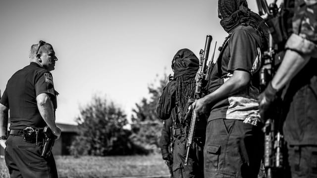 A group of armed individuals and police officer in black and white outdoor setting.